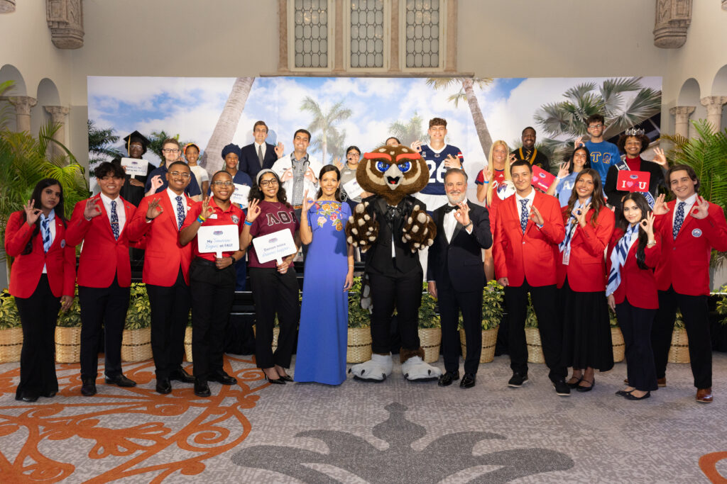group of adults stand with a mascot of an owl while some adults touch pointer finger to thumb