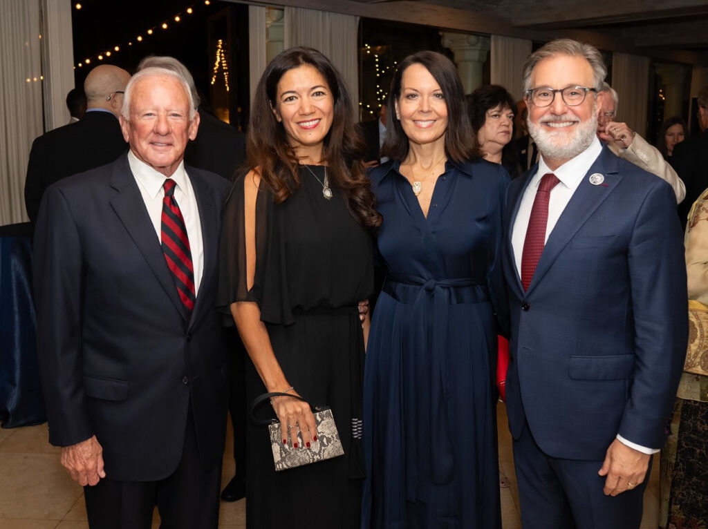 two men in suits stand with two women in dresses while smiling at the camera