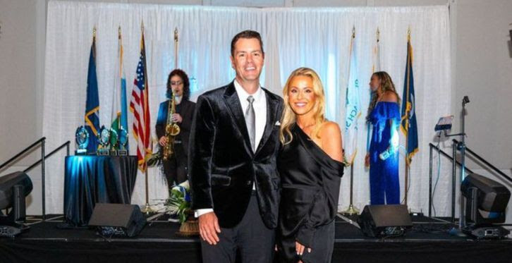 Man in black suit stands next to woman in black dress in front of a stage