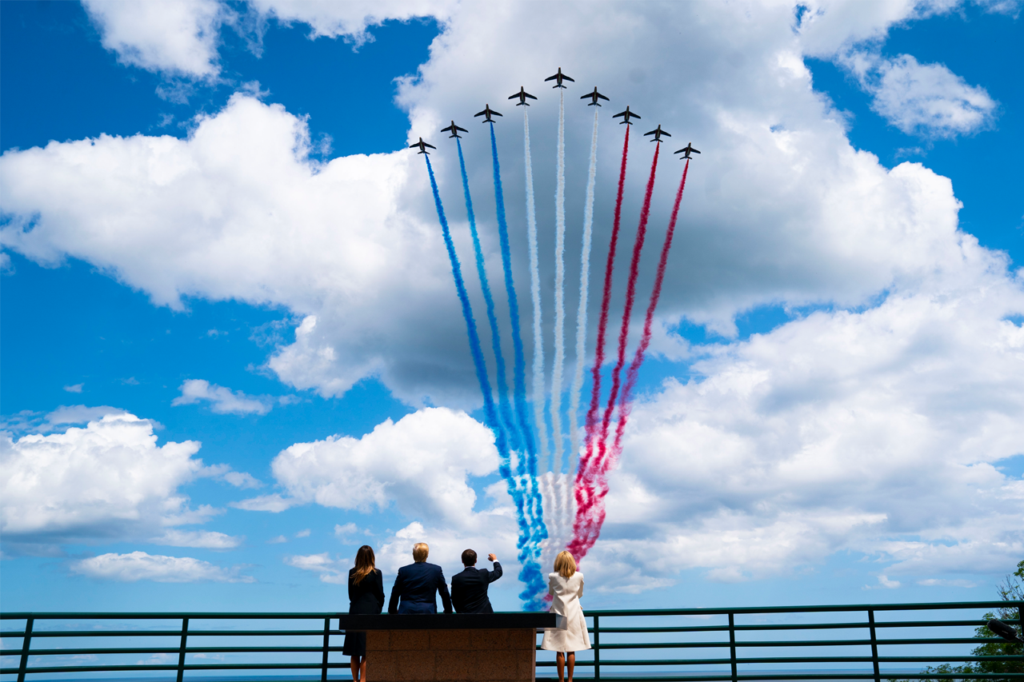 four adults stand watching planes in the sky with red white and blue smoke coming from planes