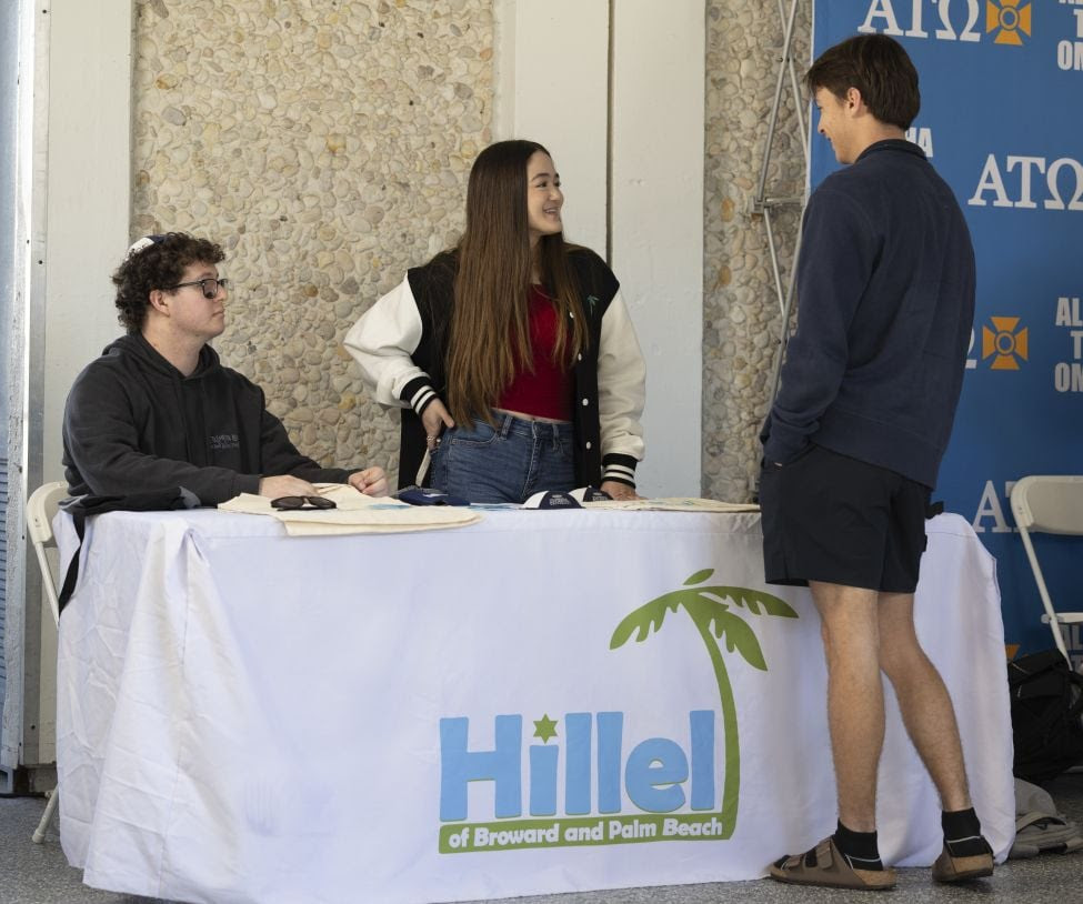 two students behind table with white table cloth talk to a student standing on other side of table