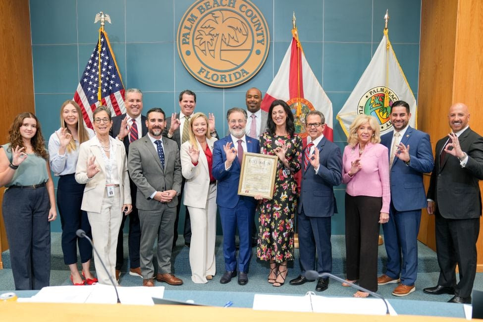 large group of adult stand with a framed paper while standing under a seal that reads "palm beach county folorida"